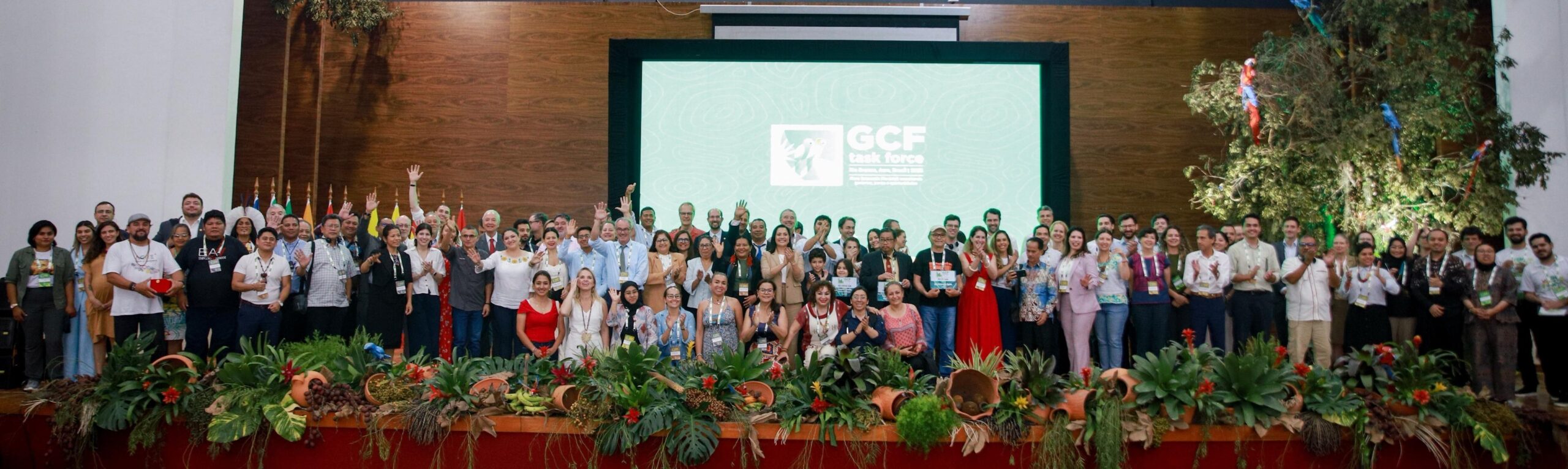 Group of people on a stage, standing in front of a screen with text GCF Task Force and behind plant decorations.