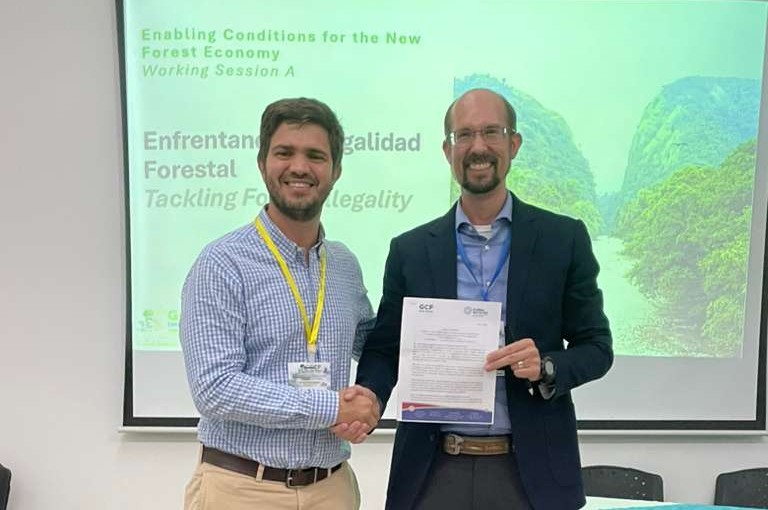 two men shaking hands in front of a screen, one is holding a document